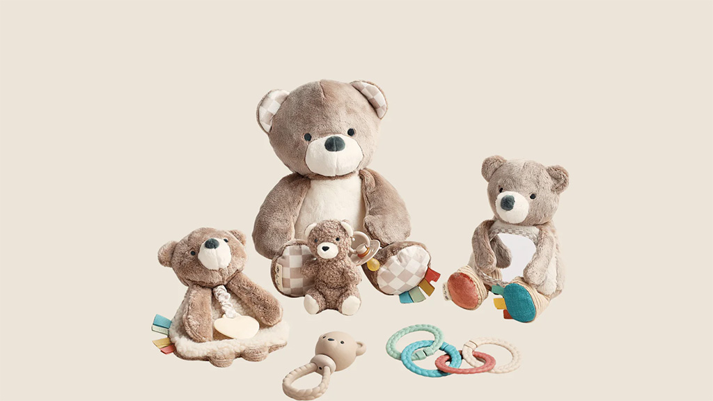 Soft plush teddy bears in various sizes arranged with pastel-colored baby rattles and teethers on a beige background.