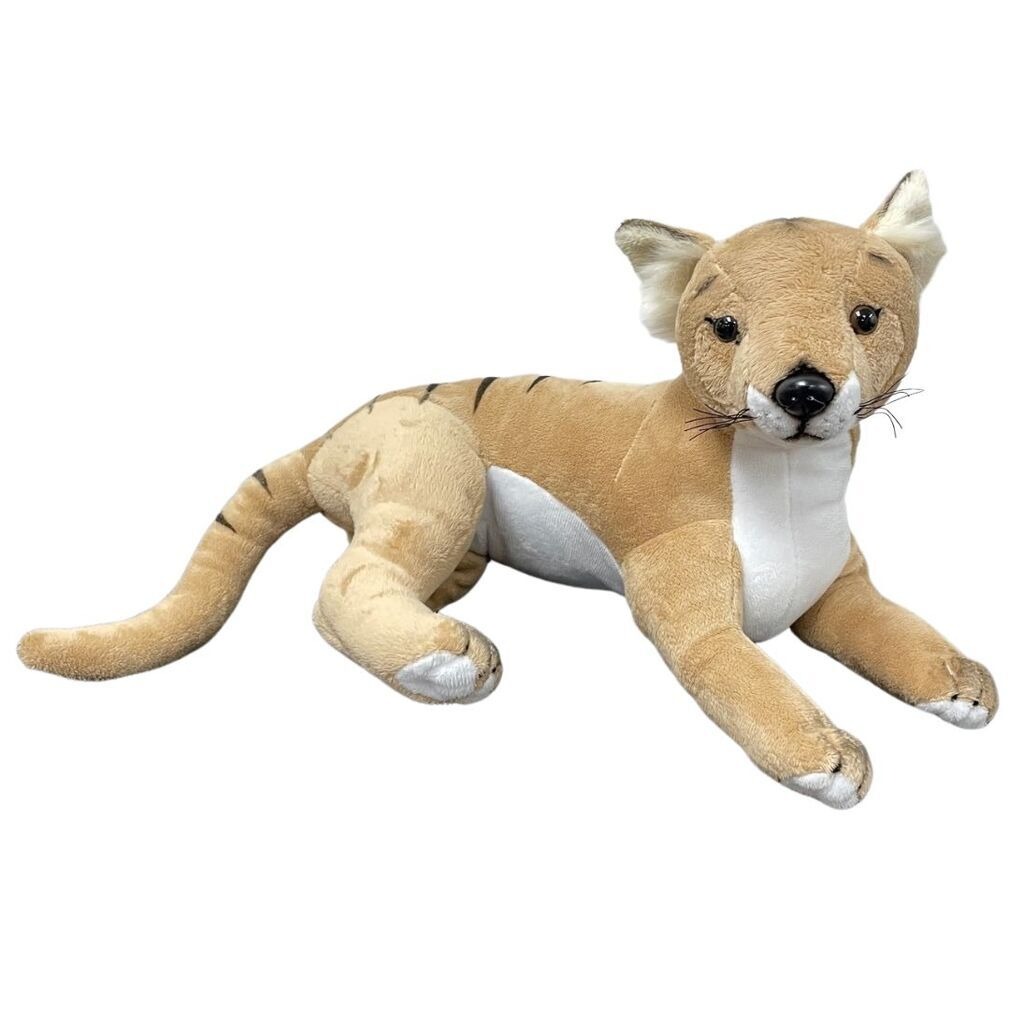 A realistic tan and white wild cat plush toy with black whiskers, pointed ears, and a long tail, lying in a stretched-out position on a white background.