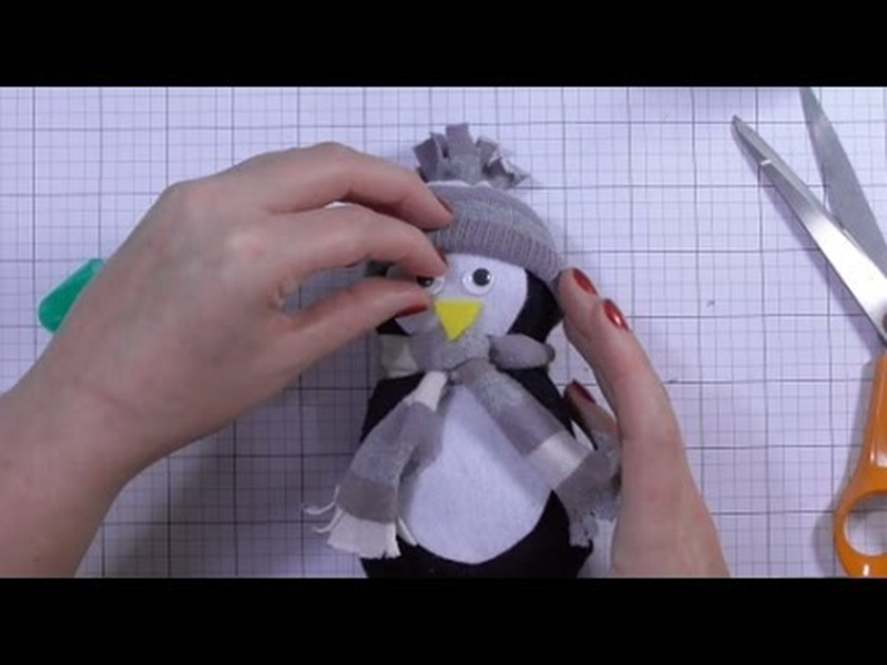 Hands assembling a small DIY penguin plush toy, adding facial details and accessories on a crafting table.