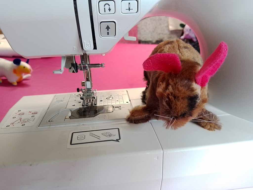 Plush rabbit toy with bright pink ears sitting on a sewing machine next to the needle area.