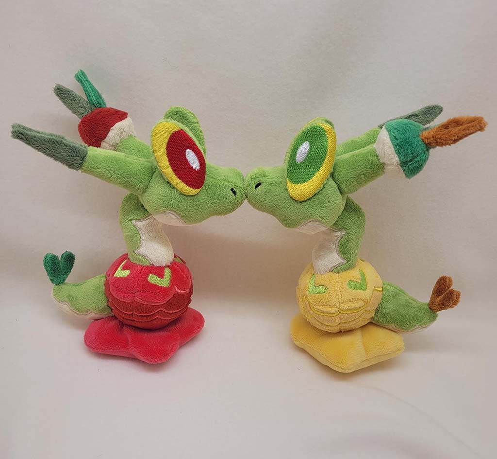 Two green dragon plush toys with large colorful eyes touch noses while standing on red and yellow embroidered bases against a neutral background.