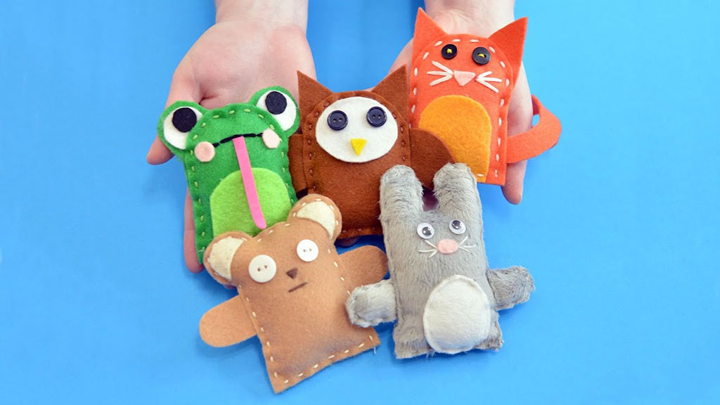 A group of small handmade felt plush animals, including a frog, owl, cat, bear, and bunny, held in two hands against a blue background, showing simple stitching and button eyes.
