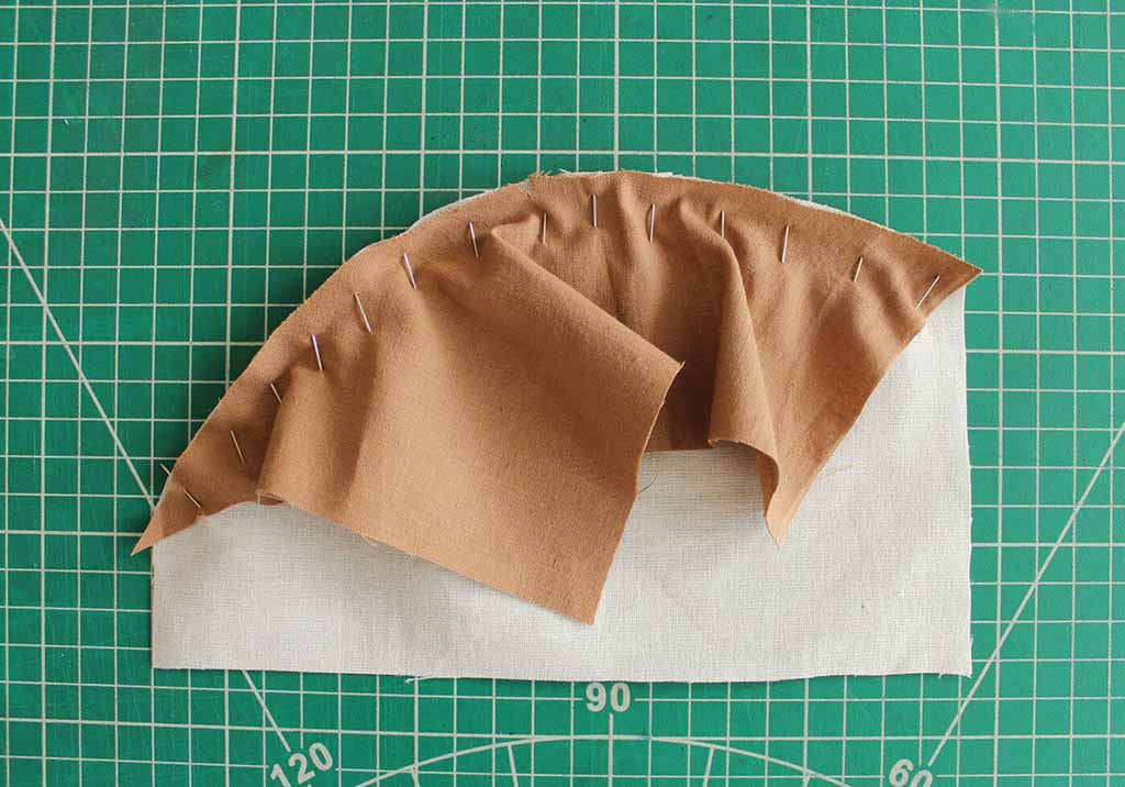 Pinned brown fabric being shaped along a curved edge on a green cutting mat, demonstrating how to align and ease fabric for sewing curved seams in plush toy making.