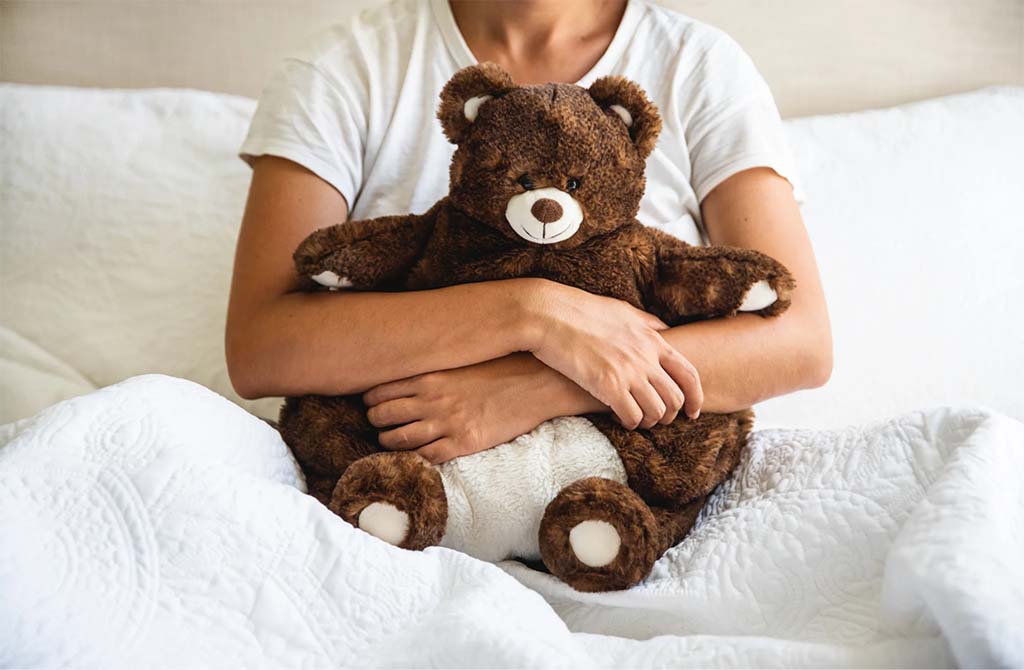 A person in a white shirt sits in bed and gently hugs a plush brown teddy bear wrapped in white blankets.