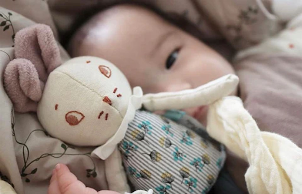 A baby lying on a bed cuddling a handmade fabric bunny plush with embroidered face and patterned clothes.