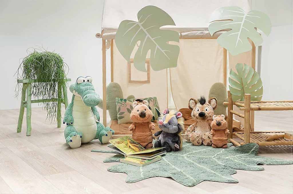 A group of soft jungle-themed plush toys—including a crocodile, hyena, hippo, and other animals—arranged in a playful indoor setup with leaf-shaped décor and natural wooden props.