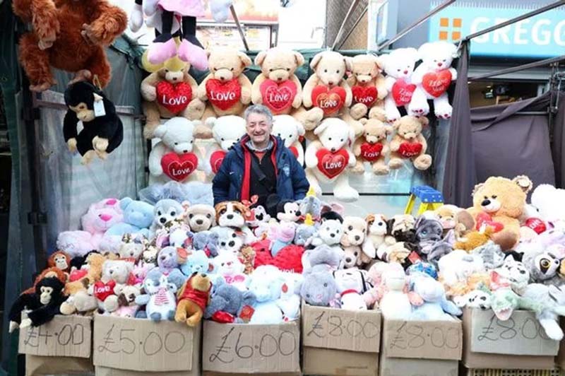 A street market vendor displaying a large collection of stuffed animals in boxes and hanging plush toys behind him.