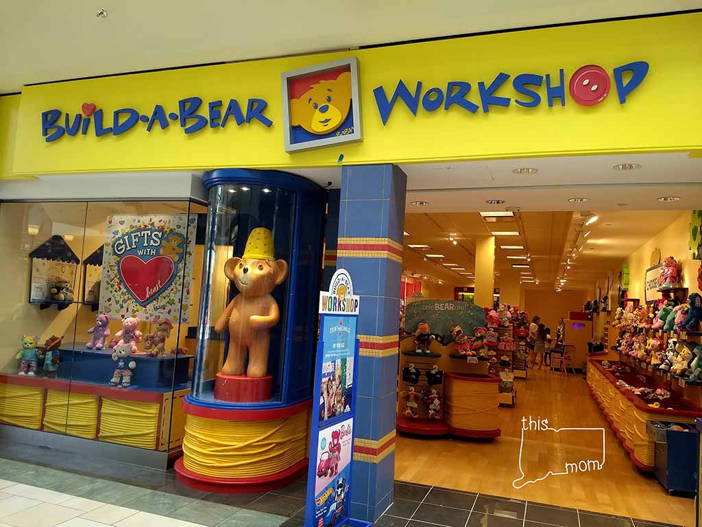 A brightly colored Build-A-Bear Workshop store entrance in a shopping mall, featuring a large bear statue, gift displays, and shelves filled with plush toys inside the store.