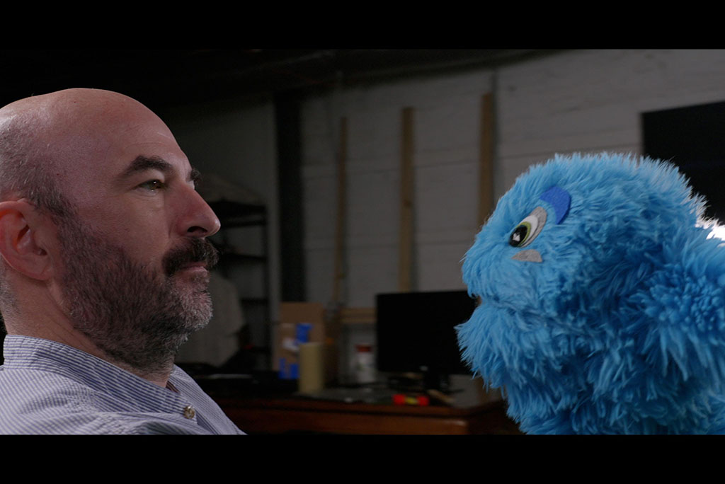 Man interacting face to face with a bright blue furry plush puppet in a studio setting.
