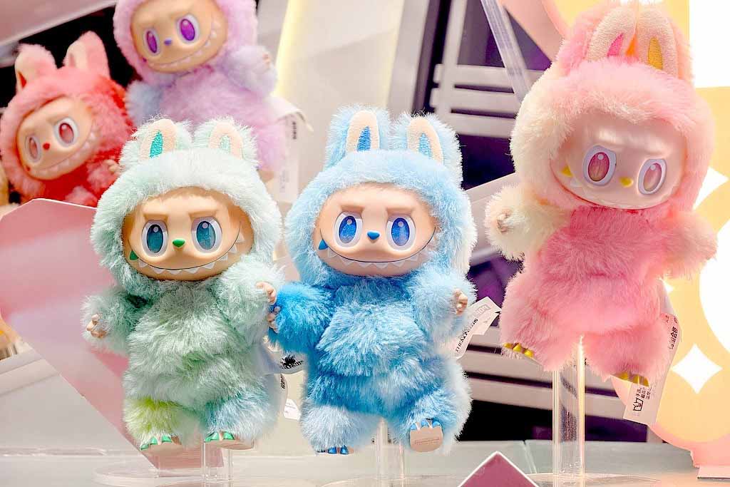 Colorful Labubu plush toys in pastel furry costumes displayed on stands in a brightly lit collectible toy shop.