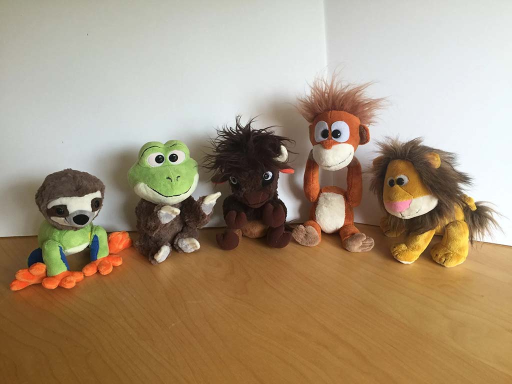 Five small plush animals—including a sloth, frog, buffalo, monkey, and lion—sitting in a row on a wooden surface against a white background.