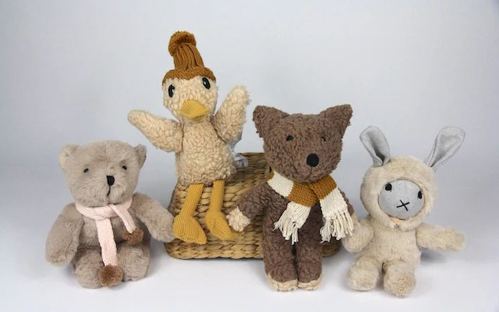 A group of handmade plush animal toys—including a beige bear, a knitted duck, a brown teddy bear with a scarf, and a soft bunny—arranged together in front of a woven basket on a light background, showing cozy textured fabrics and neutral colors.