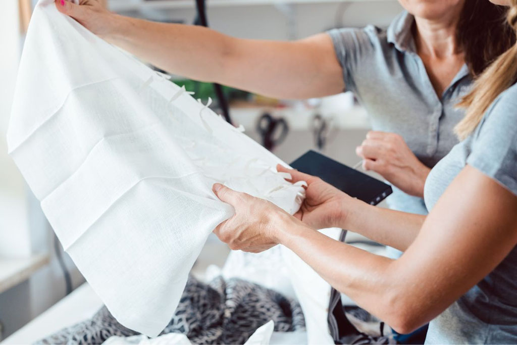 Women examining a white fabric sample closely in a workshop setting.