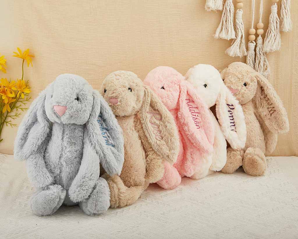 A row of soft bunny plush toys in pastel colors with embroidered names on their long ears, displayed on a light fabric background with yellow flowers and tassel decor.
