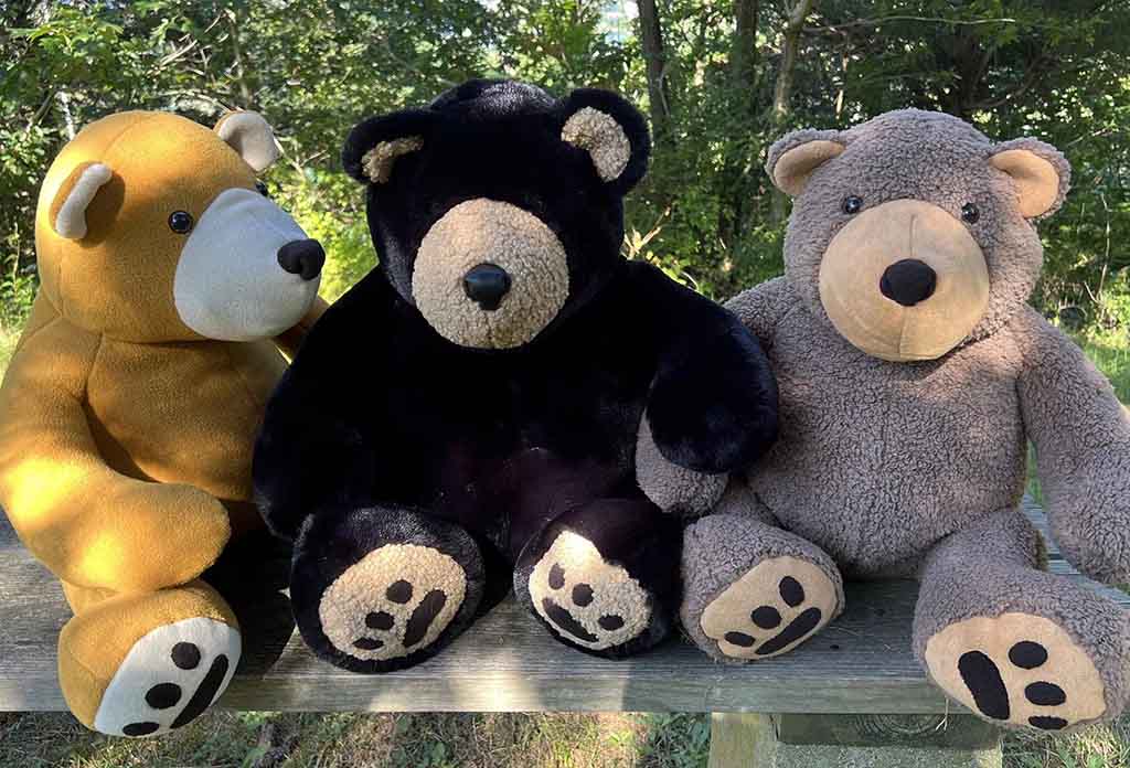 Three large plush teddy bears—one golden brown, one black, and one light brown—sit side by side on a wooden bench outdoors, with trees and greenery in the background.