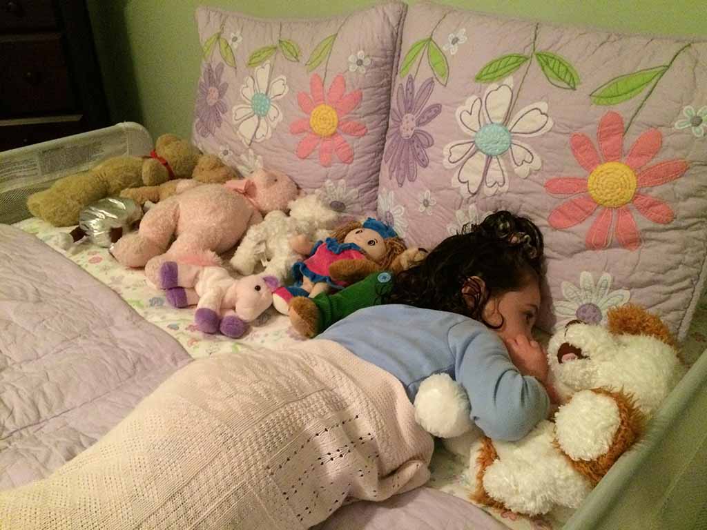 Young child sleeping in bed surrounded by multiple plush stuffed animals on floral-patterned pillows and blankets.
