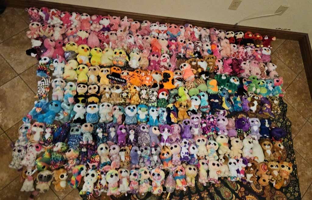 A large assortment of colorful plush toys arranged in rows on the floor, featuring various animals and characters, organized by color groups from pinks and yellows to blues and purples.
