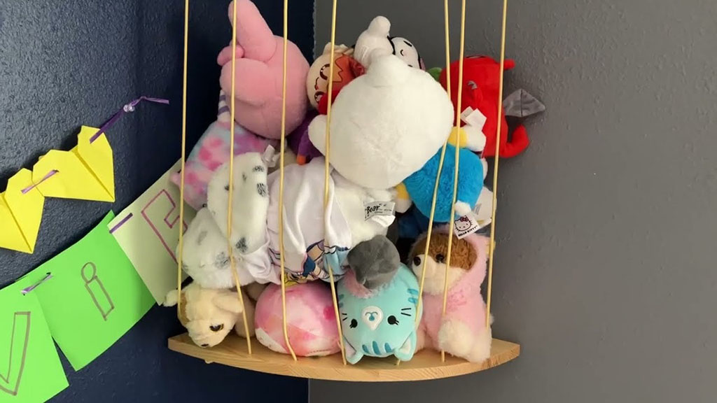 A wooden corner shelf holding a bundle of stuffed animals secured behind vertical elastic strings, organized against a dark gray wall with paper decorations hanging nearby.