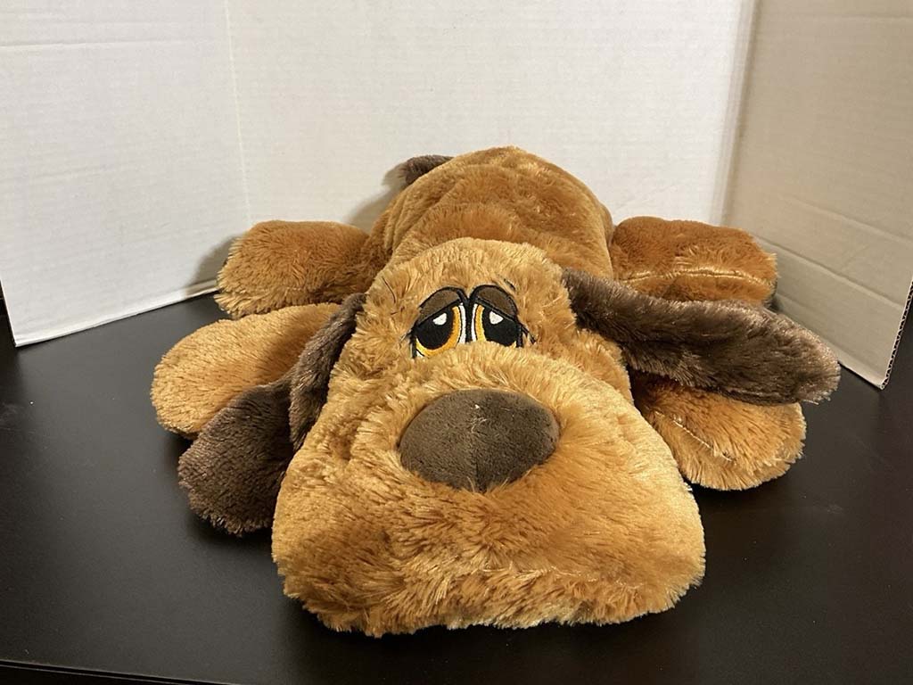 Large floppy brown plush dog lying flat on a black surface, with embroidered sleepy eyes and long droopy ears, displayed against a simple white backdrop.