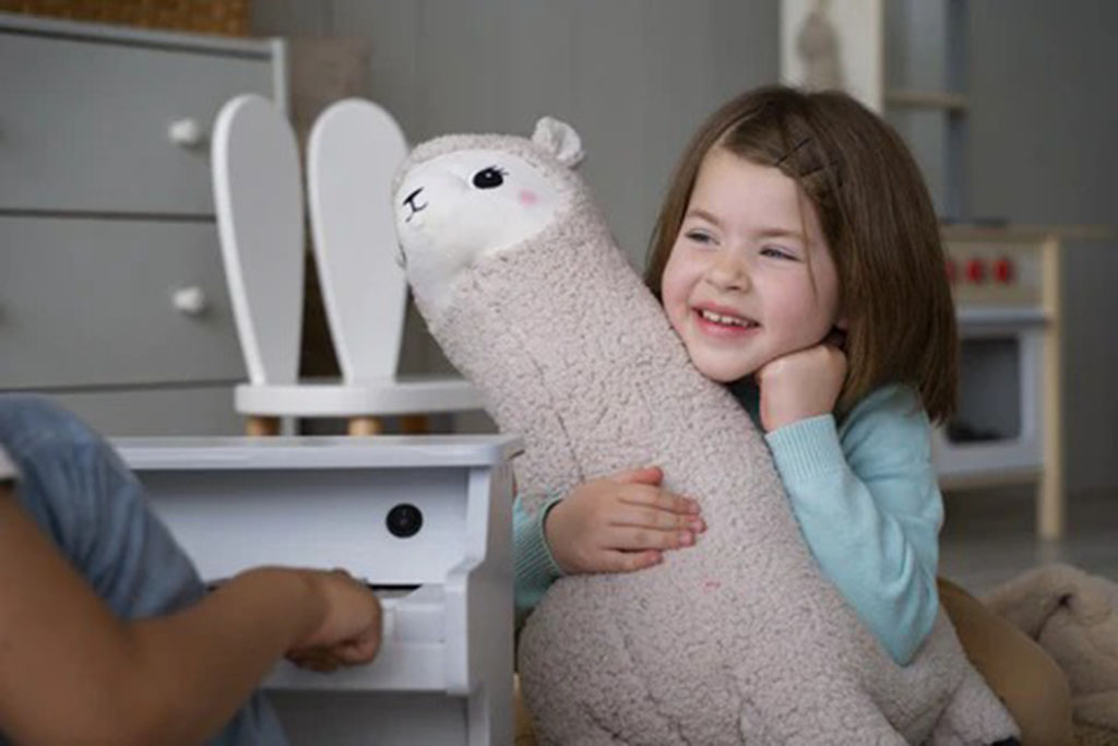 Smiling young girl sitting indoors, hugging a large soft alpaca plush toy while playing with a friend.