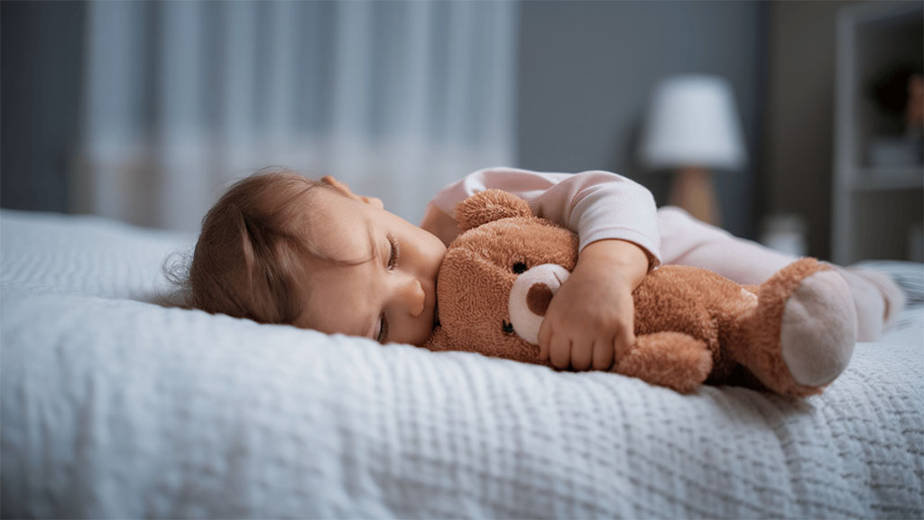 Sleeping baby cuddling a soft brown teddy bear on a cozy bed in a dimly lit bedroom.