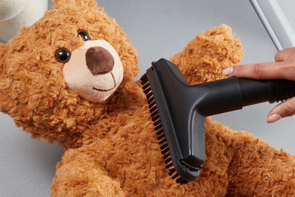 A person using a vacuum brush attachment to clean the fur of a brown teddy bear.