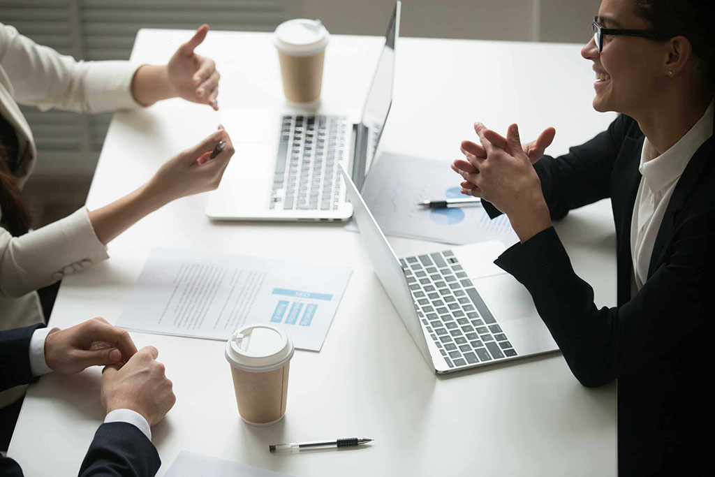 Three people in a professional meeting sitting at a white table with laptops, documents, and coffee cups while discussing business matters.