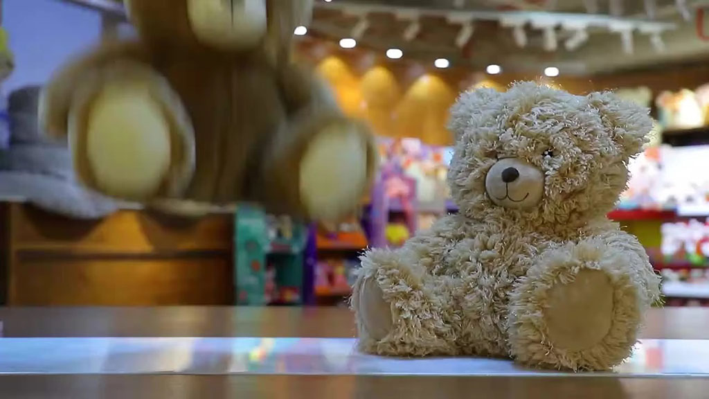 A plush teddy bear sits on a table while another teddy bear is captured mid-air during a drop test inside a brightly lit toy store.
