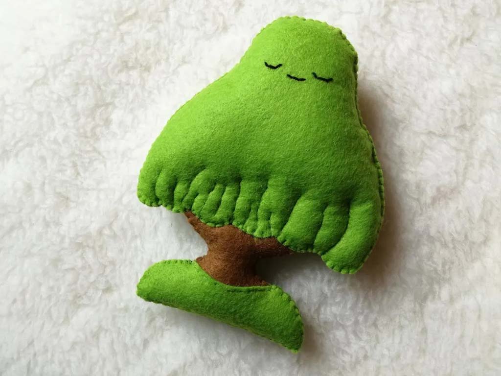 Small green felt plush toy shaped like a tree with stitched sleepy eyes, placed on a soft white fabric background.