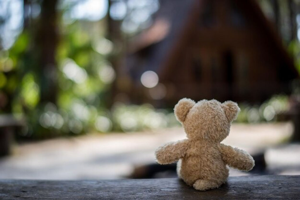 A small teddy bear sits on a wooden surface facing a blurred cabin in a forest, creating a peaceful and nostalgic outdoor scene.