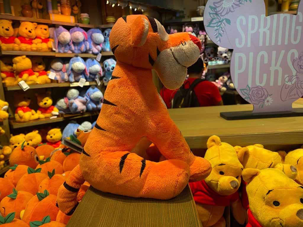 An orange Tigger plush toy displayed on a wooden shelf inside a store, surrounded by other stuffed animals like Winnie the Pooh and Eeyore, showcasing retail plush merchandising.