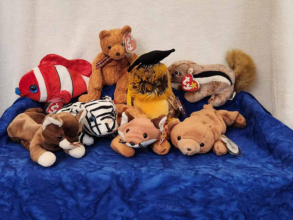 A group of Beanie Babies plush toys arranged on a blue cloth, including a clownfish, bear, owl with a graduation cap, skunk, zebra, fox, dog, and seal.