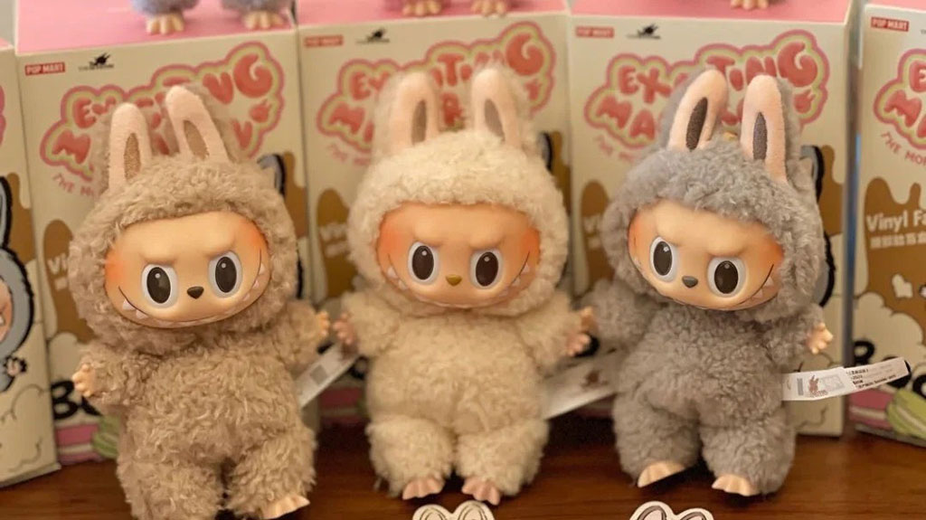 Three fluffy Labubu collectible figures dressed in soft bunny costumes in beige and gray colors, standing in front of colorful Pop Mart “Exciting Mamegoma” packaging on a display shelf.