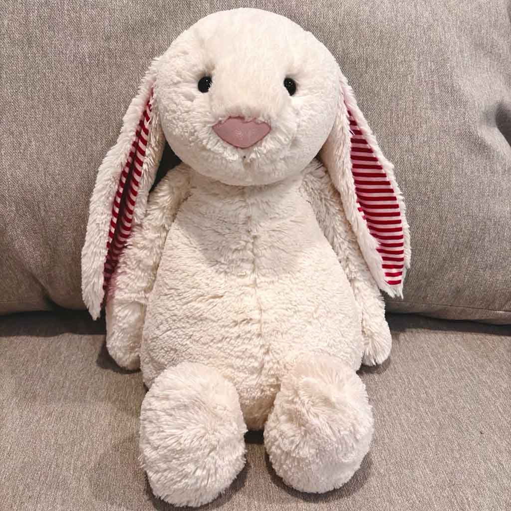 A soft white bunny plush toy with long floppy ears lined in red and white stripes, sitting on a beige sofa.