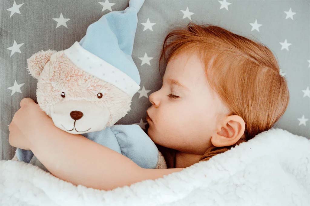 A young child with red hair peacefully sleeping while hugging a soft beige teddy bear wearing a blue nightcap and matching outfit, lying on star-patterned bedding under a cozy white blanket.