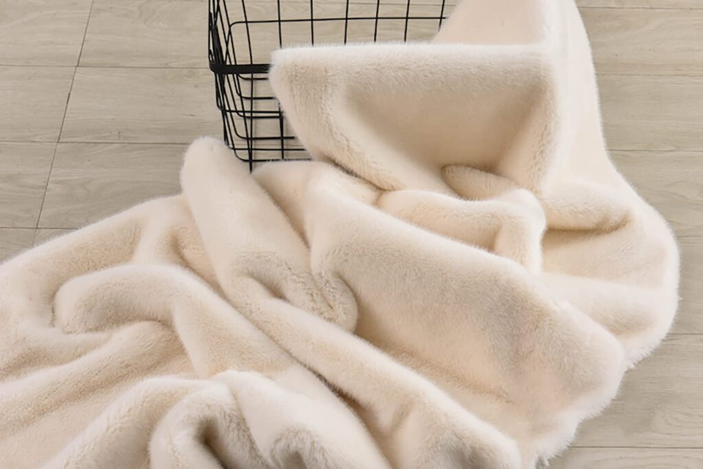 A luxurious cream-colored plush fabric blanket draped beside a black wire basket on a wooden floor, showcasing its smooth, velvety texture and cozy appearance.