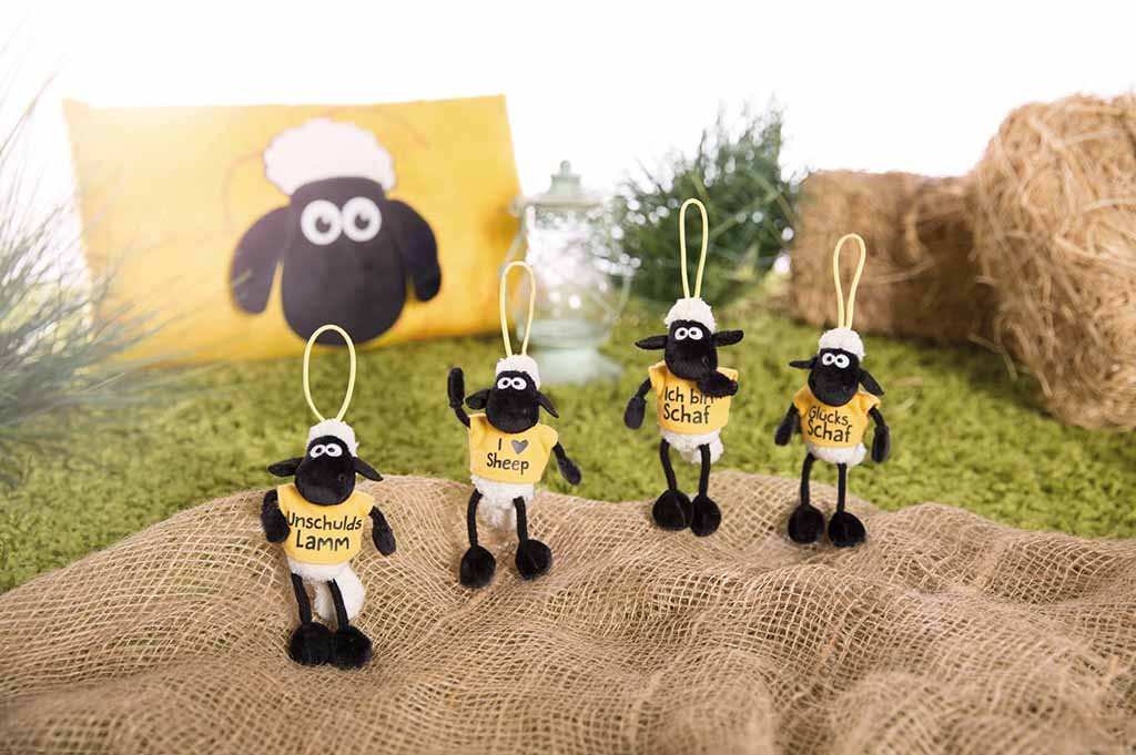 Four small sheep plush charms with yellow shirts displaying playful messages, standing on a rustic burlap cloth with green grass, hay bundles, and a sheep-themed pillow in the background.