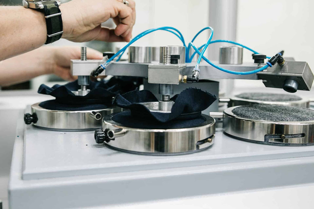 Technician operating a laboratory abrasion resistance testing machine with dark fabric samples secured under metal clamps for durability testing.