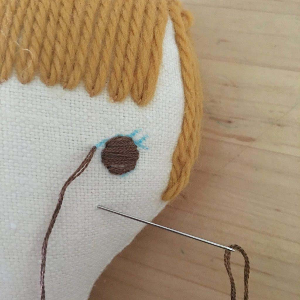 Close-up of a handmade plush doll face being embroidered, showing brown thread stitching an eye detail on fabric with yarn hair.