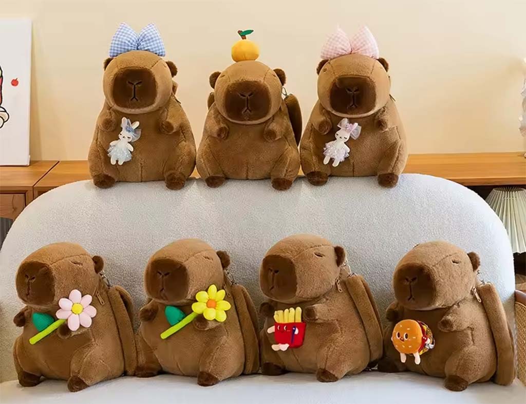 A group of soft brown capybara plush backpacks with different accessories like bows, flowers, and mini toys, displayed neatly on a couch and wooden shelf.