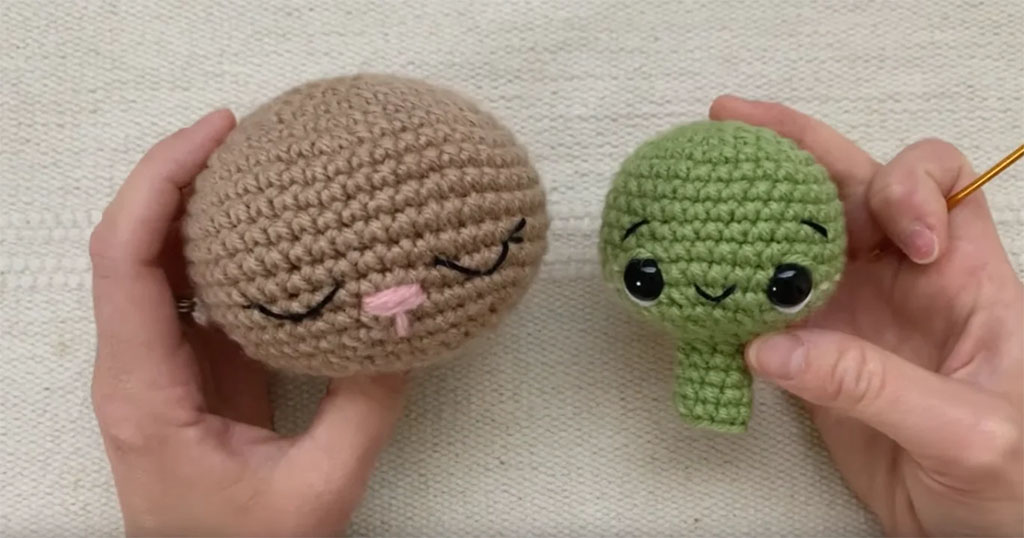 Two crocheted amigurumi heads held in hands, one beige with embroidered sleepy eyes and a pink nose, and one green with safety eyes and a small smile.