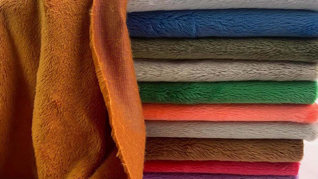 Close-up of stacked plush fabrics in various colors including orange, blue, green, gray, pink, and brown, showcasing the smooth texture and thickness used for stuffed toys and plush manufacturing.