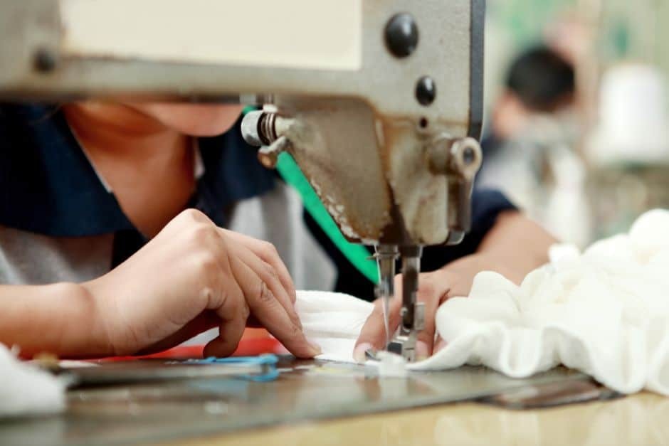 Close-up of a worker using an industrial sewing machine to stitch white fabric, showing hands guiding the material during plush toy production.