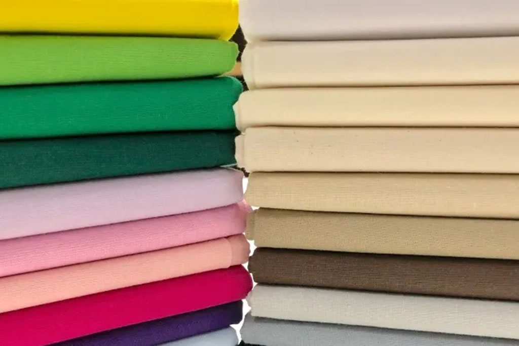 Stacks of neatly folded cotton fabrics in a wide range of colors, from bright greens and yellows to soft pastels and neutral tones.