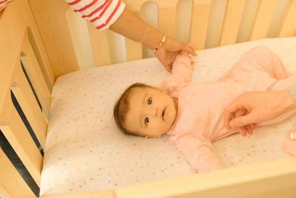 Baby lying on a crib mattress in a soft pink outfit while caregivers gently hold her arms, ensuring comfort and safety.