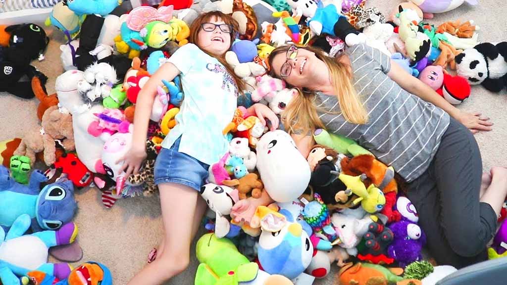 A smiling woman and child lie on a carpet surrounded by a large colorful pile of stuffed animals and plush toys, creating a playful and joyful scene.