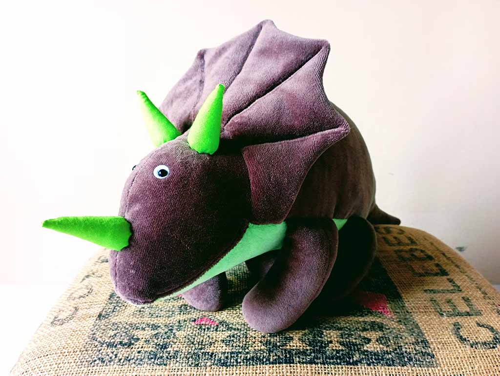 A soft triceratops plush toy with dark brown fabric, bright green horns, and a matching green belly, displayed on a textured burlap-style surface.
