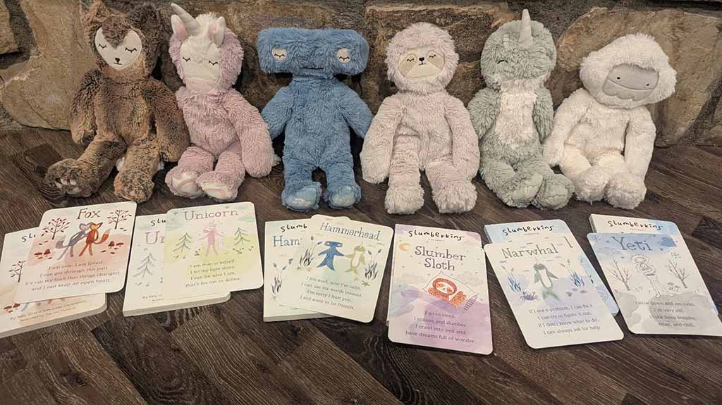 A row of Slumberkins plush creatures—including a fox, unicorn, hammerhead, sloth, narwhal, and yeti—displayed on a wooden floor with their matching affirmation story cards arranged in front of them.