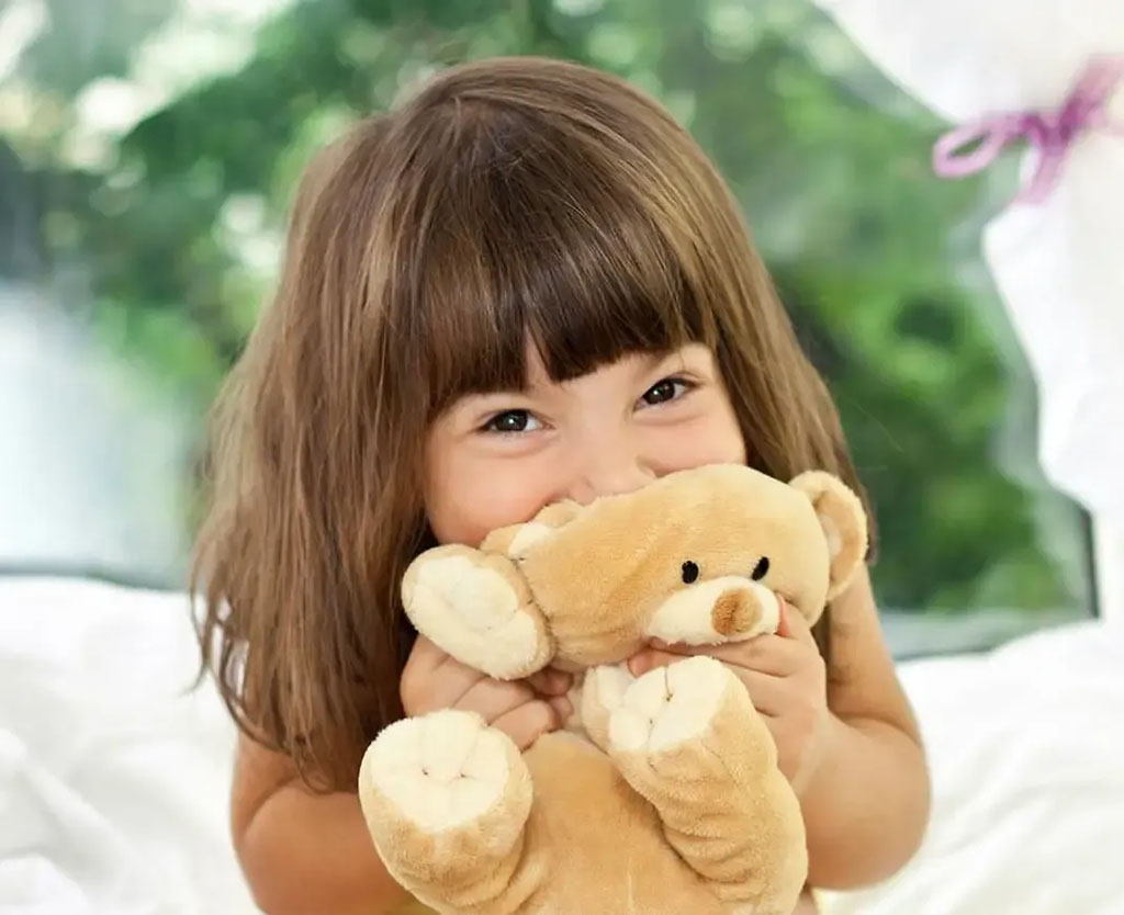 A young child with brown hair smiling joyfully while hugging a soft beige teddy bear indoors with greenery visible in the background.
