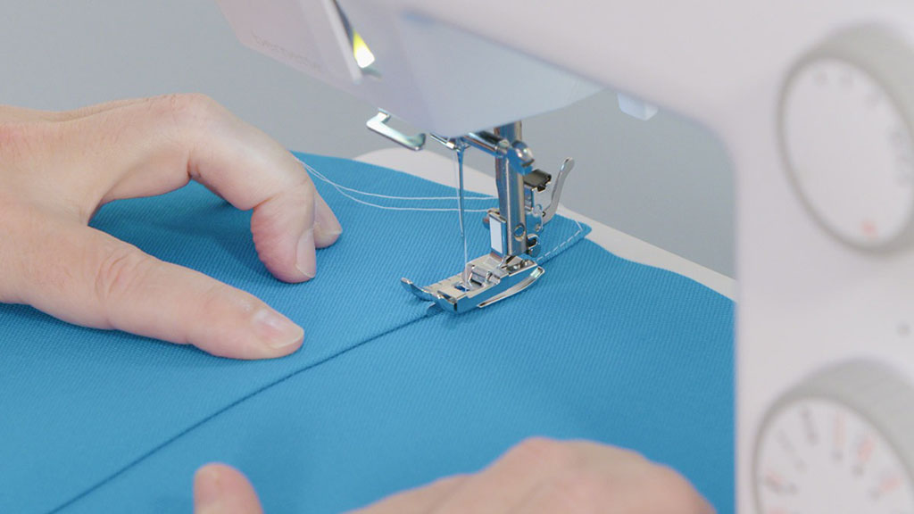 Close-up of hands guiding blue fabric through a sewing machine, showing detailed stitching and precise edge sewing technique.